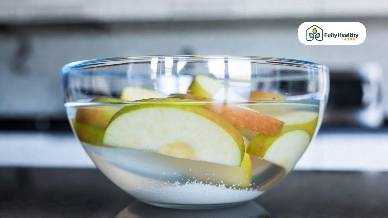 Apple slices soaking in a bowl of water with salt to prevent browning on a kitchen counter.