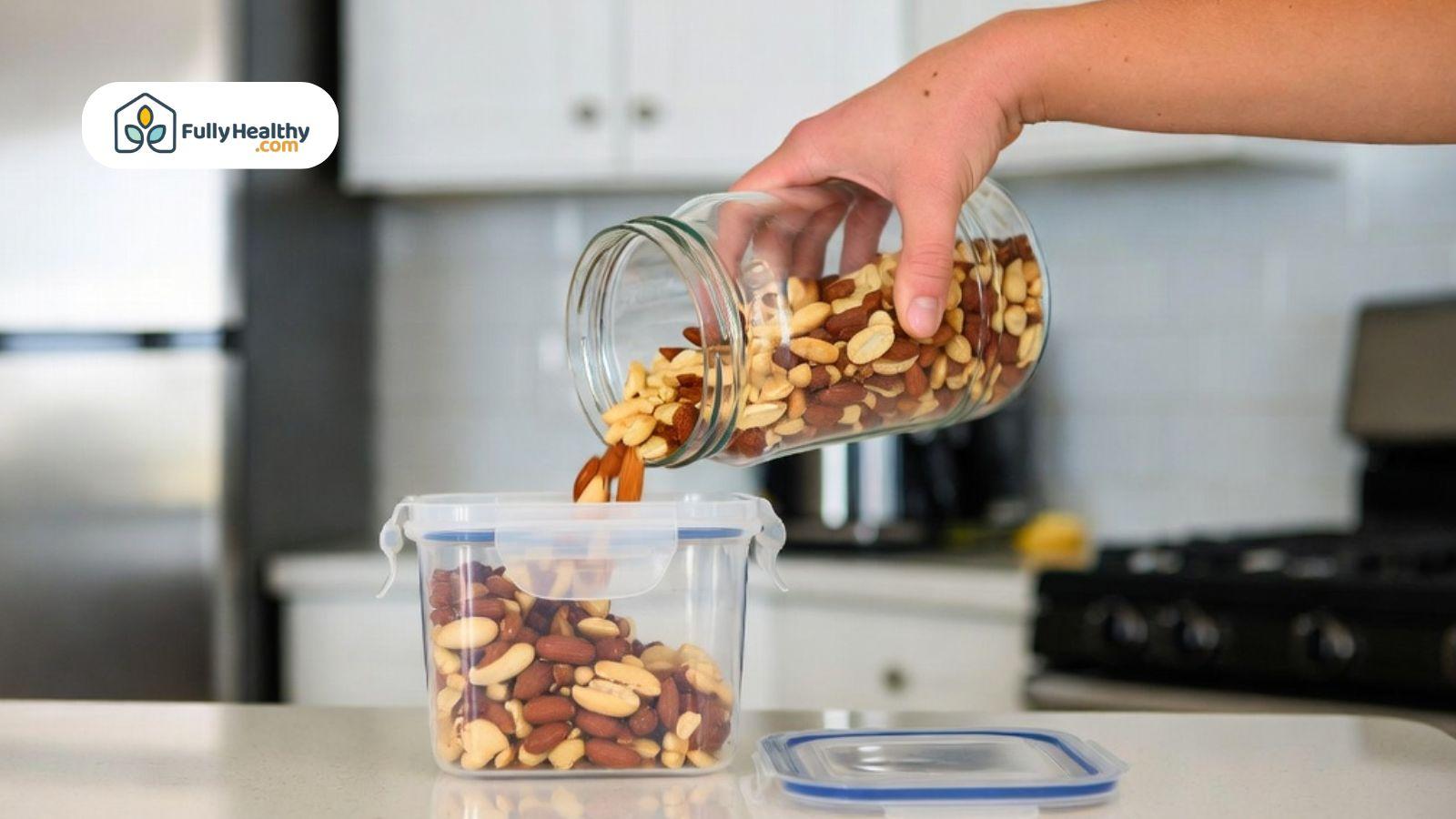 Hand pouring mixed nuts from a glass jar into a plastic airtight container in a kitchen.
