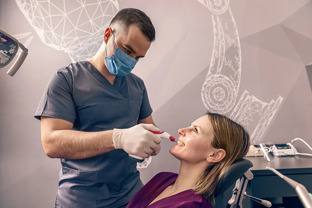 A dentist in scrubs and mask treating a smiling patient in a dental chair with decorative wall art visible in the background