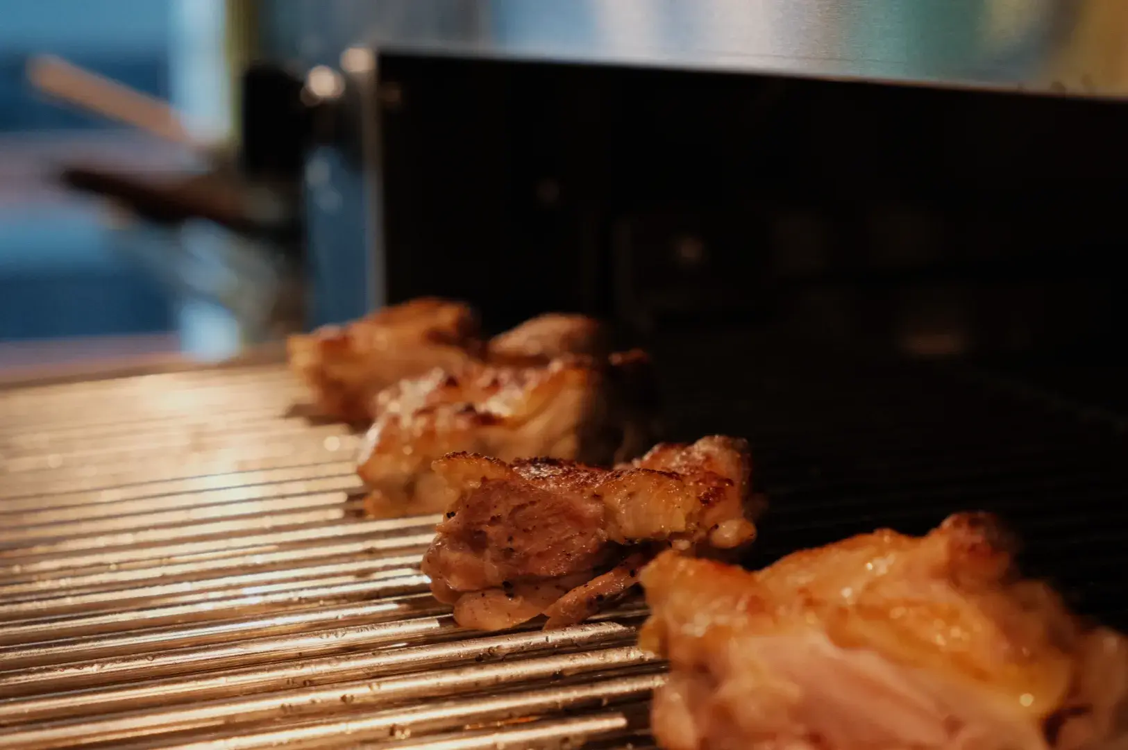 Golden-brown chicken pieces roast on a metal grill beneath a dark oven hood, with heat and savory aromas suggesting a warm, appetizing kitchen scene.