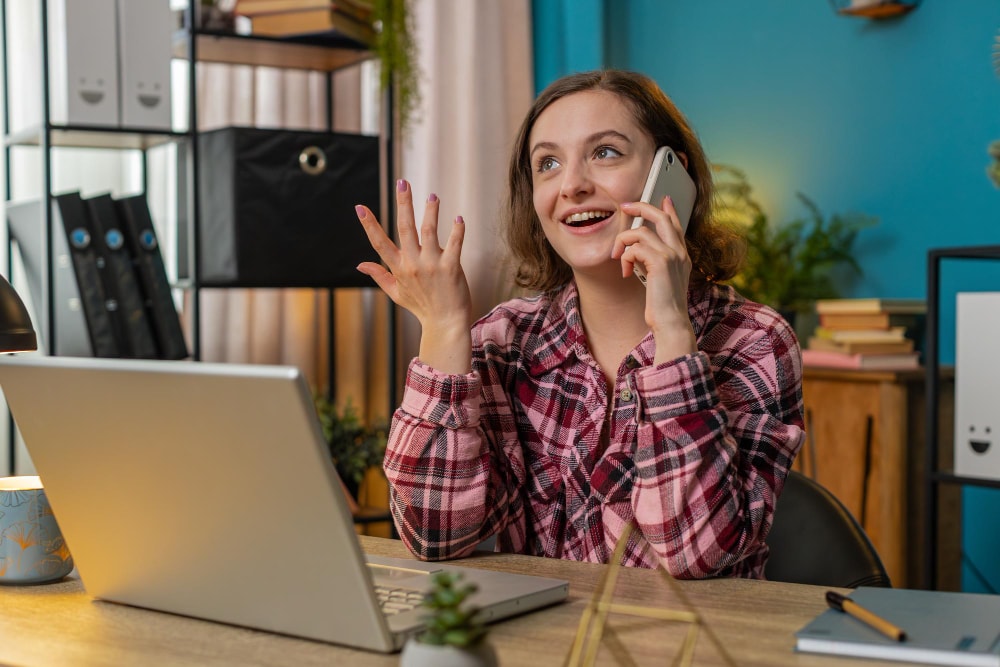 Cheerful young woman in a plaid shirt speaking on a smartphone at her home office desk, reflecting the convenience of modern business phone systems enabling seamless communication anywhere.