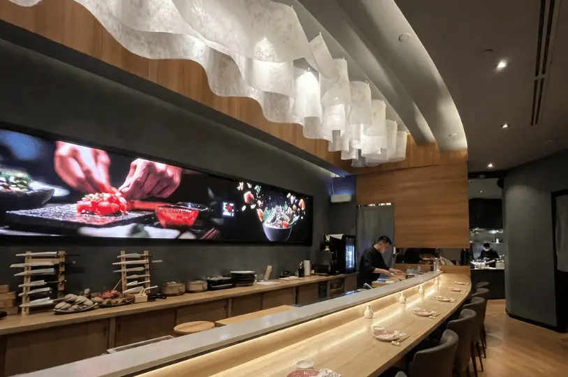Elegant and modern Japanese restaurant interior in Singapore with a curved wooden sushi bar, decorative white wavy ceiling lights, and a staff member preparing food behind the counter.
