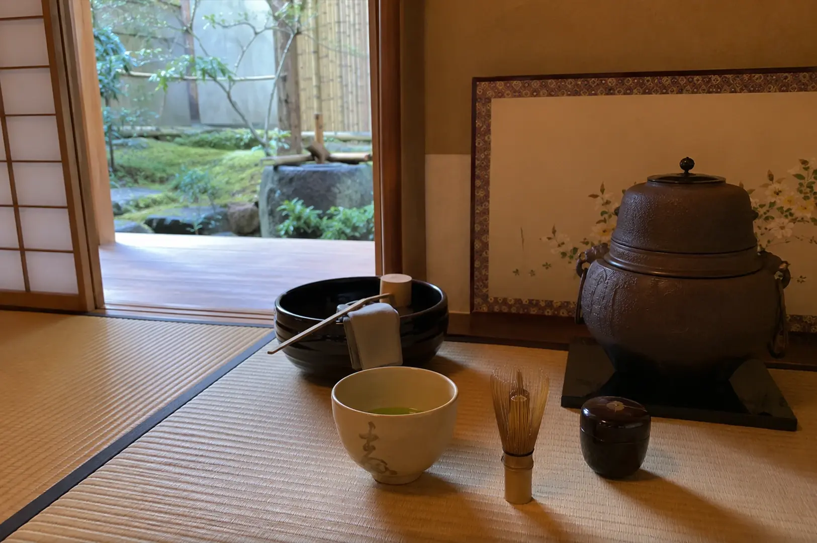 A close-up shot of a traditional Japanese tea preparation arrangement set on a woven tatami mat next to an open sliding shoji door. In the foreground, a light-colored ceramic bowl filled with vibrant green matcha tea stands beside an upright bamboo whisk and a small, polished black lacquer container. Behind them, a large dark iron kettle sits on a black platform in front of a decorative table screen painted with delicate white flowers. The open doorway in the background offers a peaceful view of a mossy outdoor garden with a stone water basin.