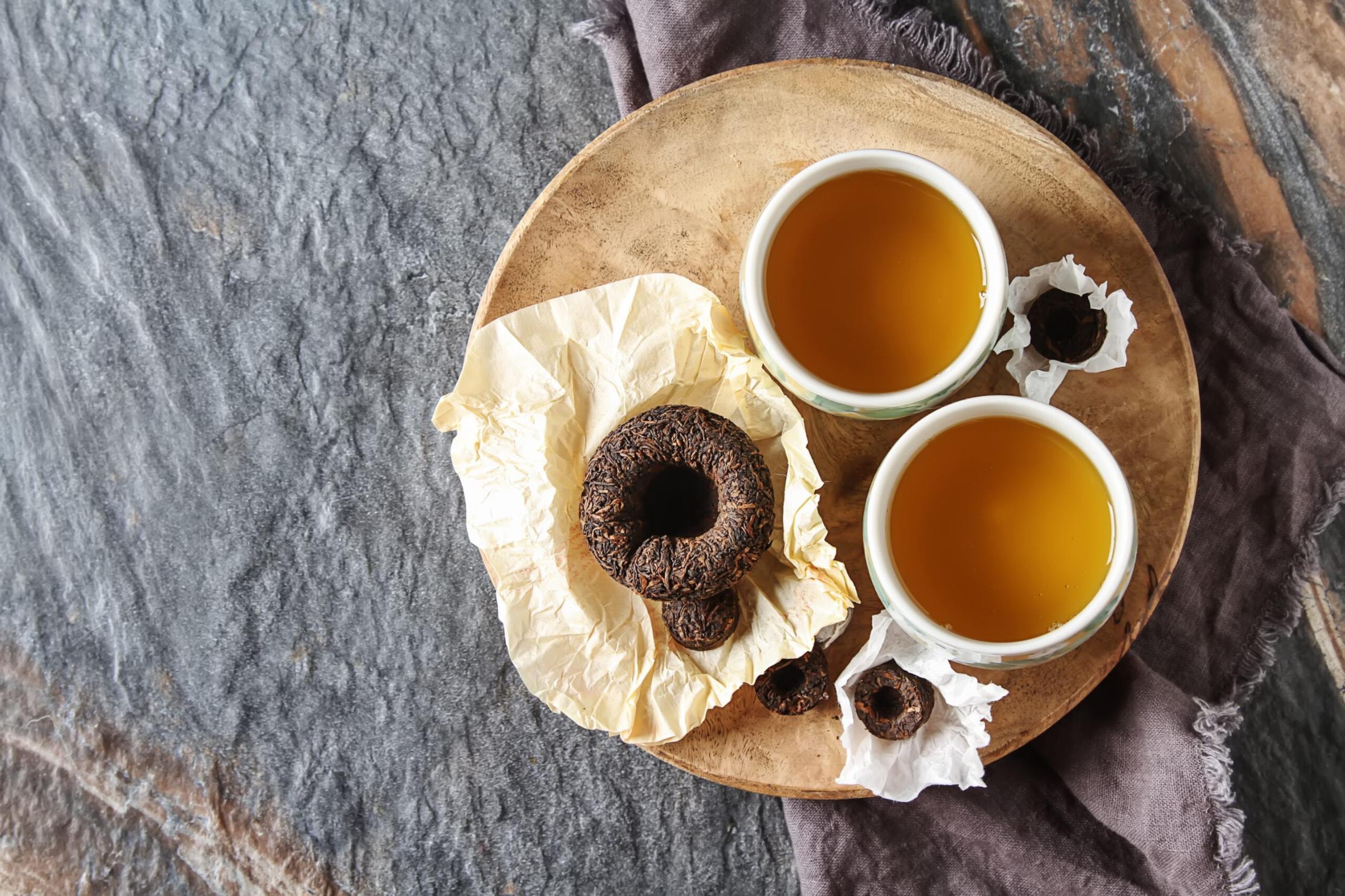 This image features two cups of golden tea and several compressed tea cakes displayed on a wooden platter over a textured stone surface. The arrangement includes both large and small tea disks resting on crinkled paper, creating a rustic and traditional aesthetic.