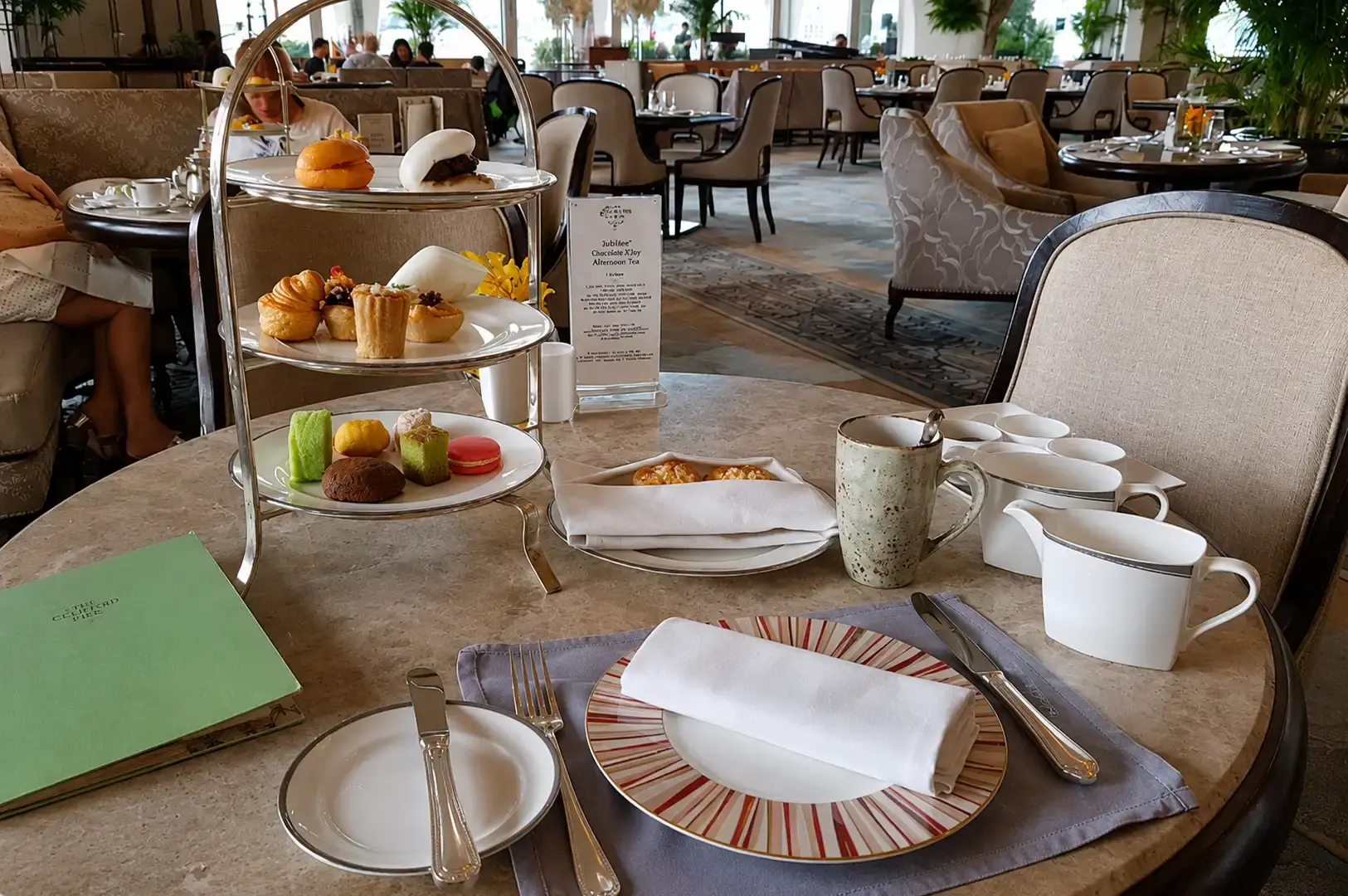 A marble tabletop fully set for high tea, featuring a three-tiered stand of savory and sweet items, a green menu, a woven napkin resting on a colorful patterned rim plate, a rustic mug, white creamer and sugar containers, and comfortable plush armchair seating in the background.
