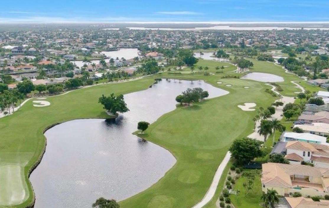 The image depicts a pristine golf course green with a water hazard, framed by luxurious homes in the background during the golden hour, showcasing the scenic beauty of Marco Island's golf course communities. The warm light enhances the vibrant greens and the elegance of the residential architecture, creating a stunning backdrop for outdoor leisure.