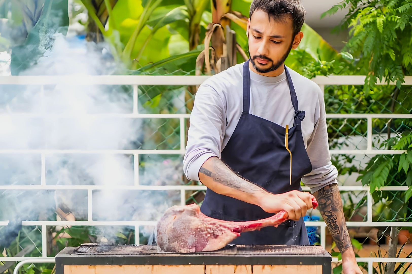 A man in an apron grills a large piece of meat outdoors, surrounded by greenery. Smoke wafts from the barbecue, creating a focused, serene atmosphere.