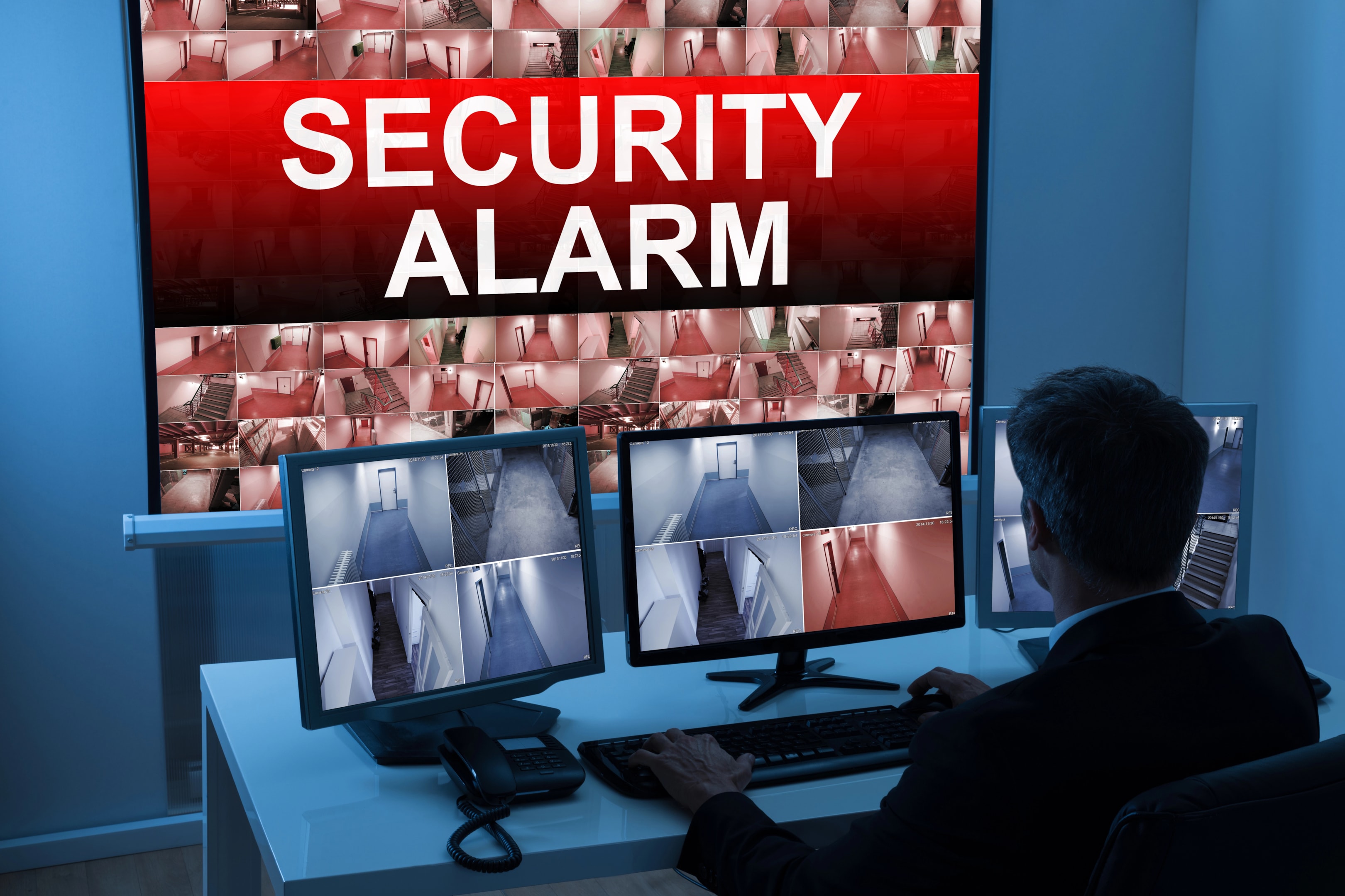 На изображението мъж наблюдава изображенията от различни камери за видеонаблюдение на екрани, докато една от камерите е активирала алармена система. Системата за видеонаблюдение осигурява сигурност на търговския обект и анализира поведението на клиентите.