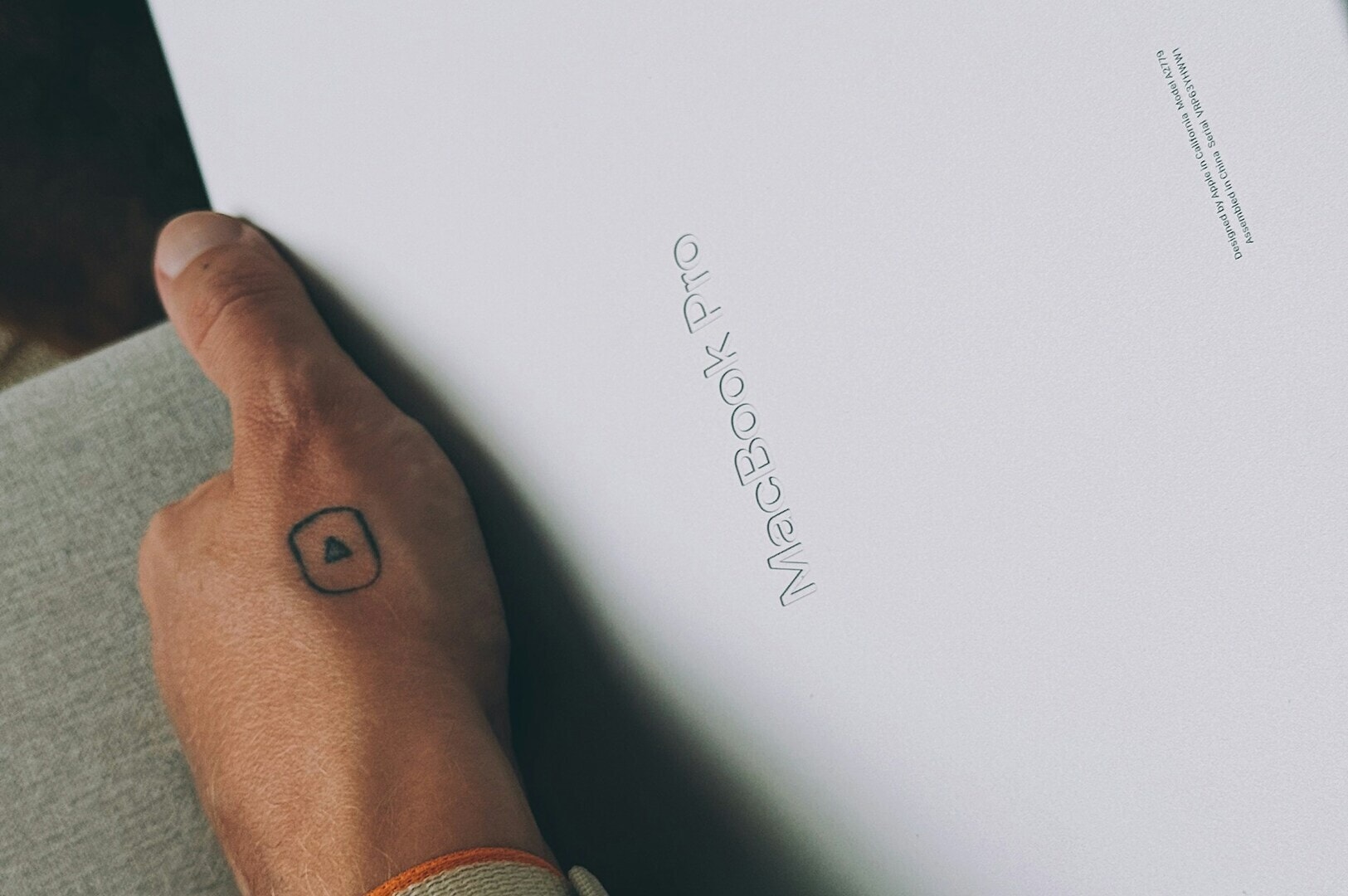 A hand with a small triangle tattoo holds a white MacBook Pro box on a sofa. The image conveys anticipation and excitement for new technology.