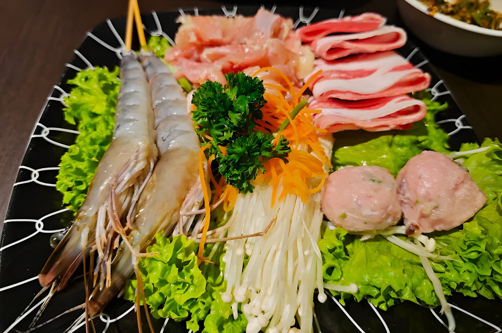 A black plate with fresh ingredients including shrimp, enoki mushrooms, sliced meat, and meatballs, garnished with lettuce, carrot, and parsley.