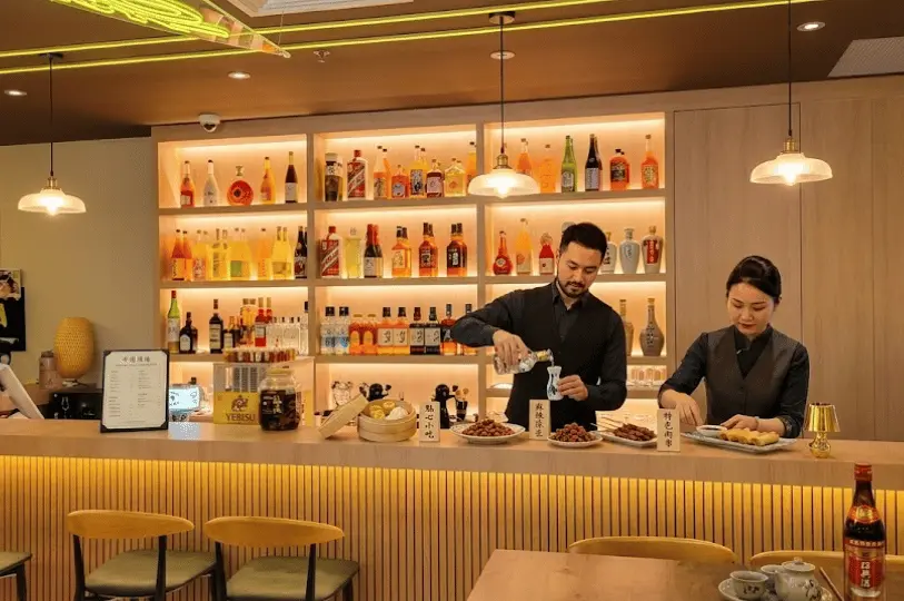 A modern, warmly lit restaurant bar where a bartender pours a drink and a staff member prepares spring rolls, with a backlit display of alcohol bottles in the background.