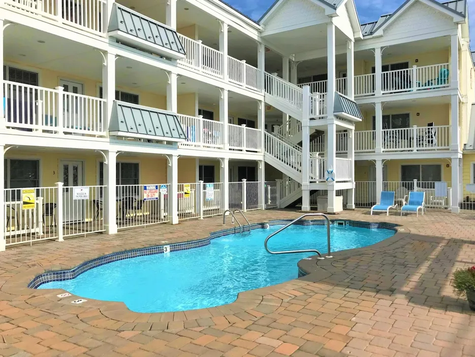 View of the Summers Haven complex in Lavallette, New Jersey, featuring a private in-ground pool with lounge chairs, brick patio, and three-story building with white railings and balconies.