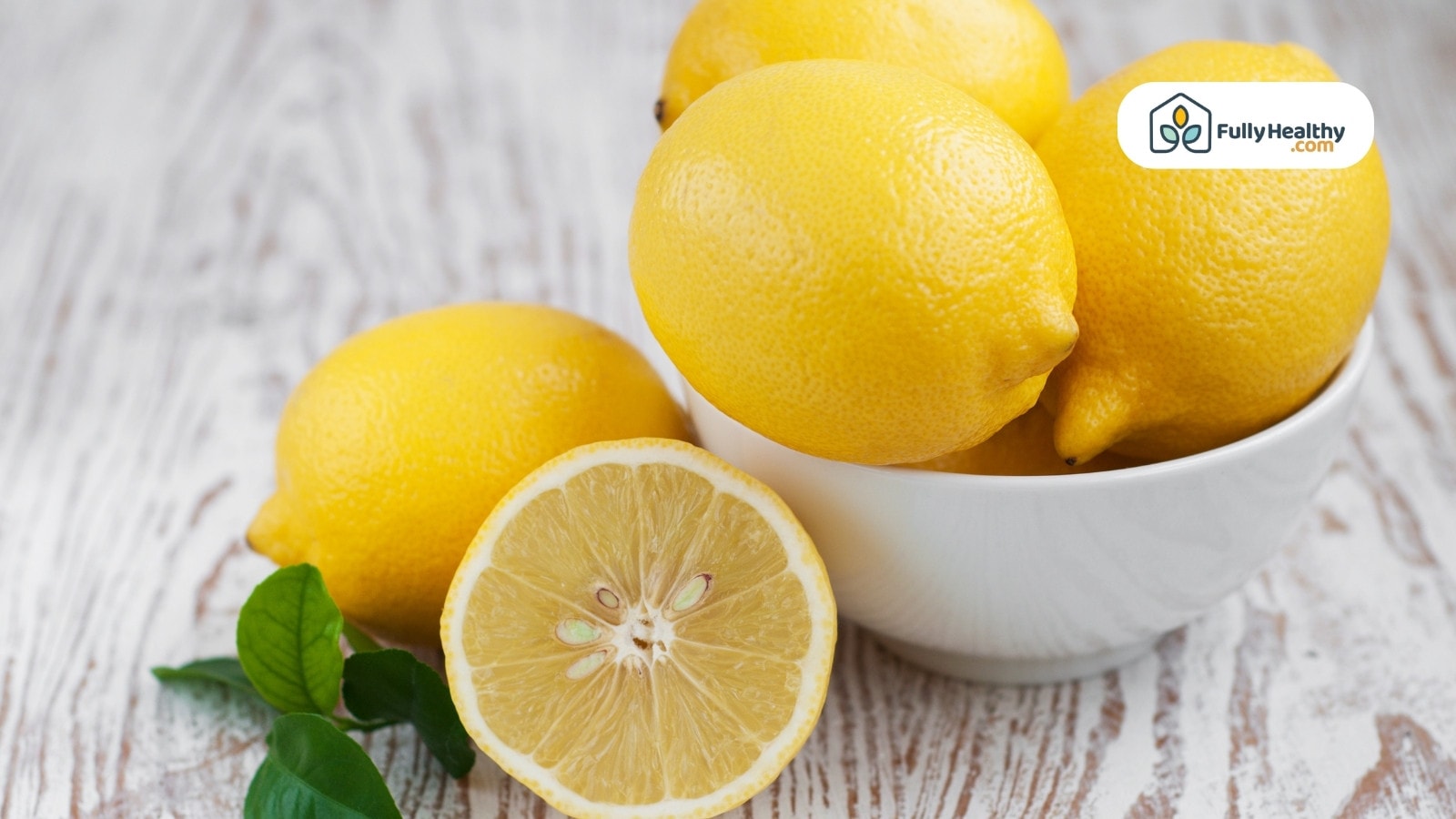 Whole and halved lemons in a white bowl with green leaves