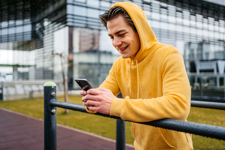 Young man in a yellow hoodie leaning against a railing sending a text.  
