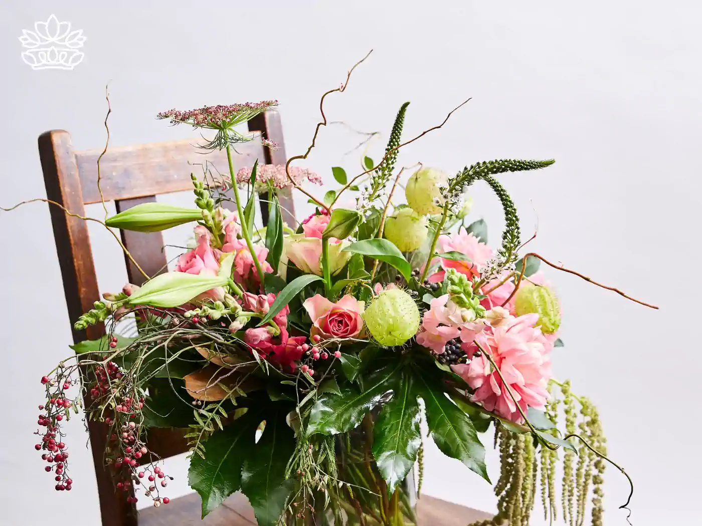 Charming arrangement of pink roses mixed with delicate blush snapdragons and greenery, gracefully presented in a clear vase on a vintage wooden chair.