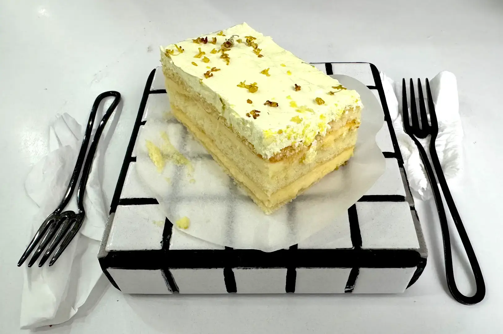 A slice of layered cake topped with yellow frosting and edible flowers, served on a 2D grid-patterned ceramic stand with black cutlery at Cafe Monochrome.