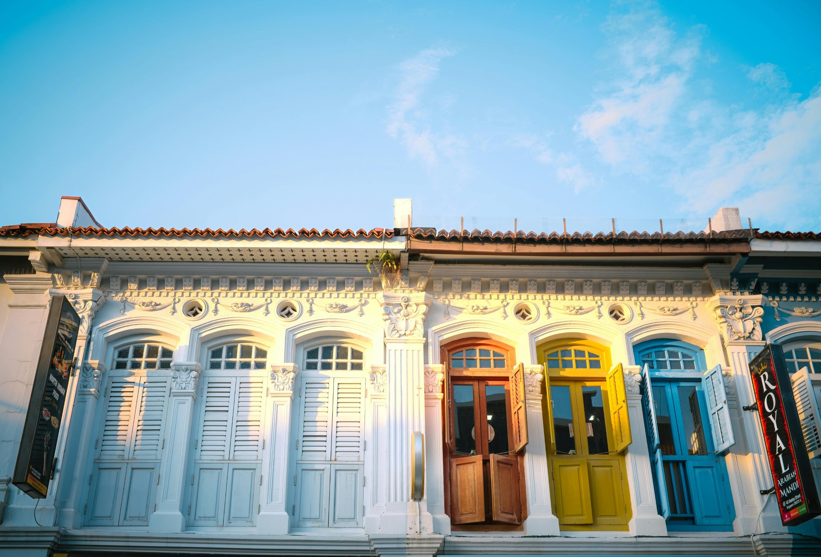  This image captures the upper story of a white colonial-style building featuring ornate architectural details and a series of arched windows with classic shutters. Three of the windows are painted in bold, contrasting colors of orange, yellow, and blue, standing out vividly against the white facade under a clear, bright sky.