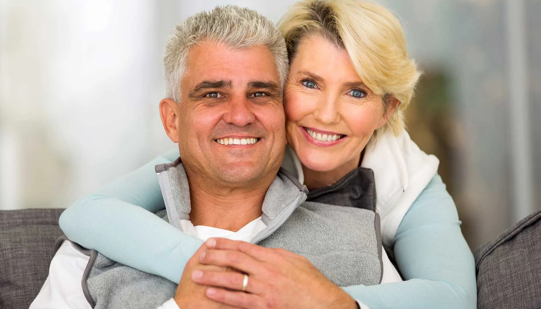 All-on-4: Tannløsningen Som Forandrer Livet Ditt På Én Dag 13 Smilende eldre par som sitter tett sammen i sofaen, begge ser glade og avslappede ut med hvite tenner og varme uttrykk.