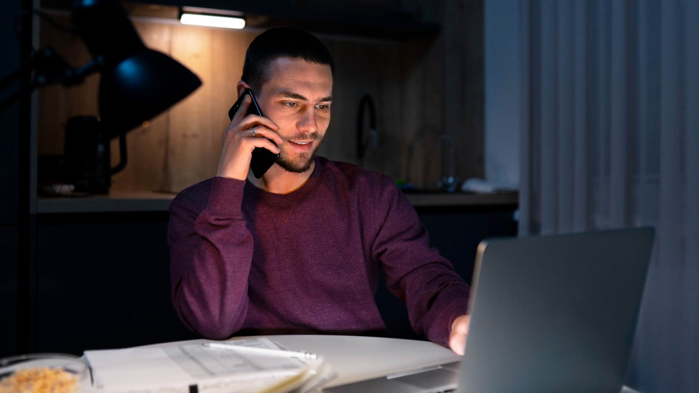 Man working late at a kitchen table, on a phone call while using a laptop, reflecting the role of security in cloud-based phone systems.