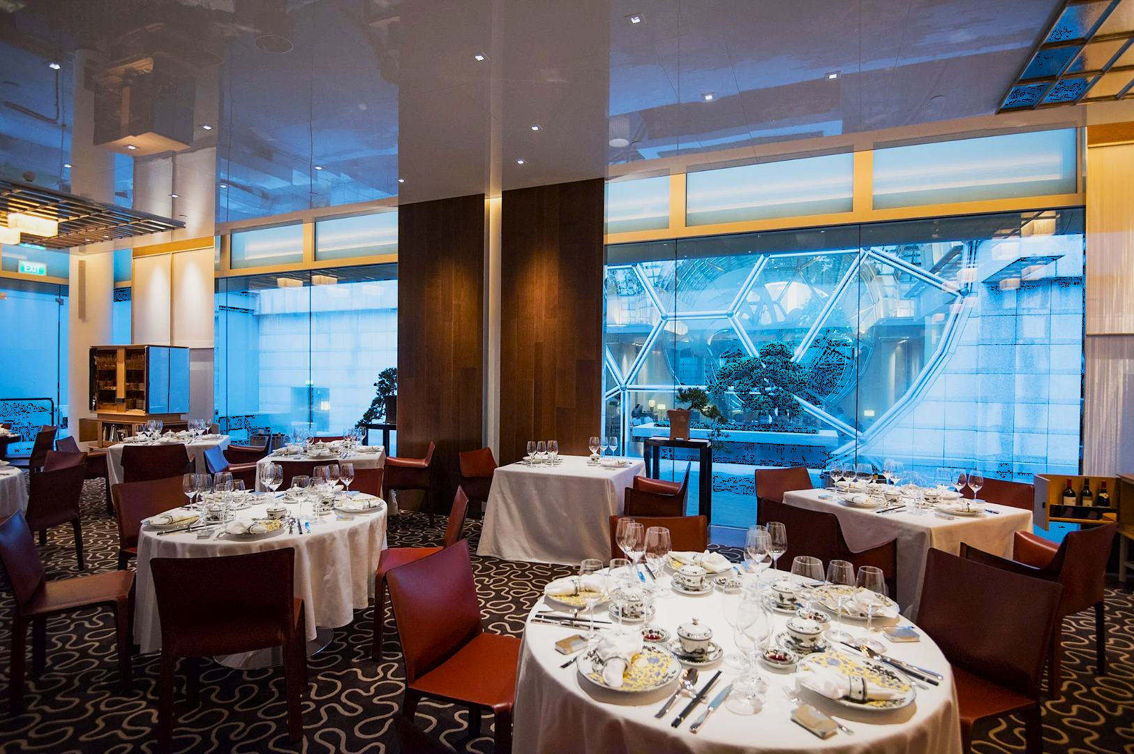 Elegant restaurant interior with round, white-tablecloth tables set with fine china, glasses, and cutlery, against a modern glass wall. Ambient lighting creates a sophisticated atmosphere.