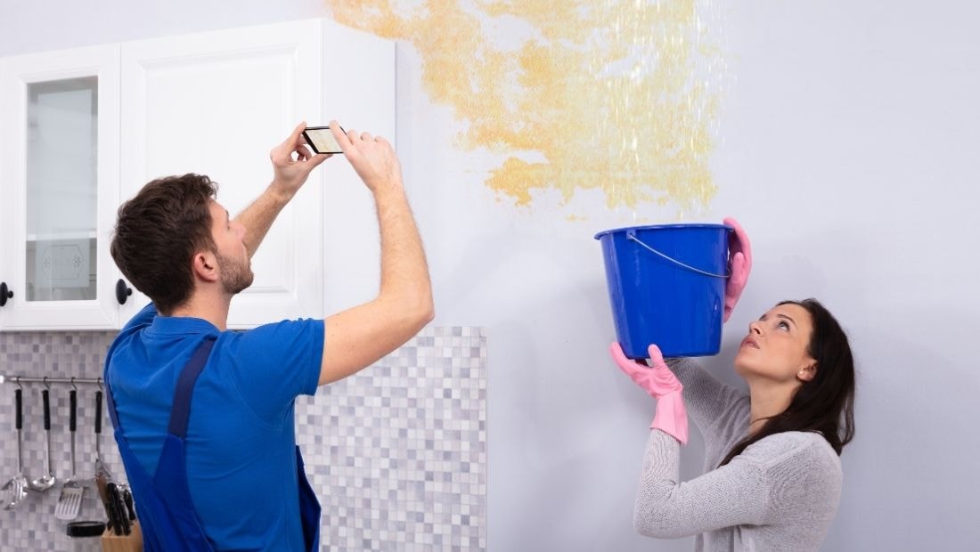 A woman holding a blue bucket while looking up at water damage on walls, as a man takes a photo of the damage. Both are wearing gloves and working together on a home repair project.