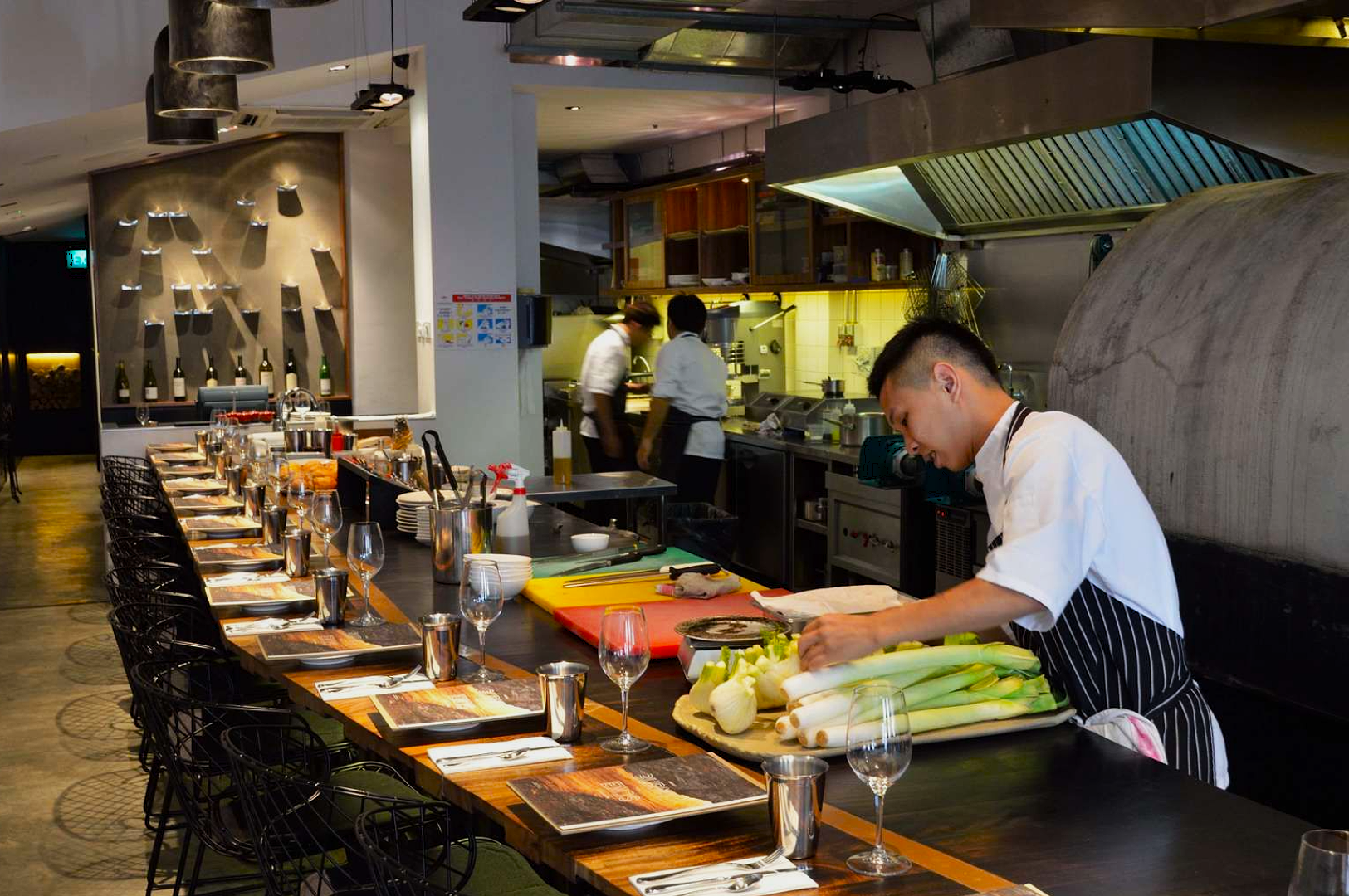 A chef prepares vegetables at a sleek, modern open kitchen with a long, set dining table. The atmosphere is lively and professional.