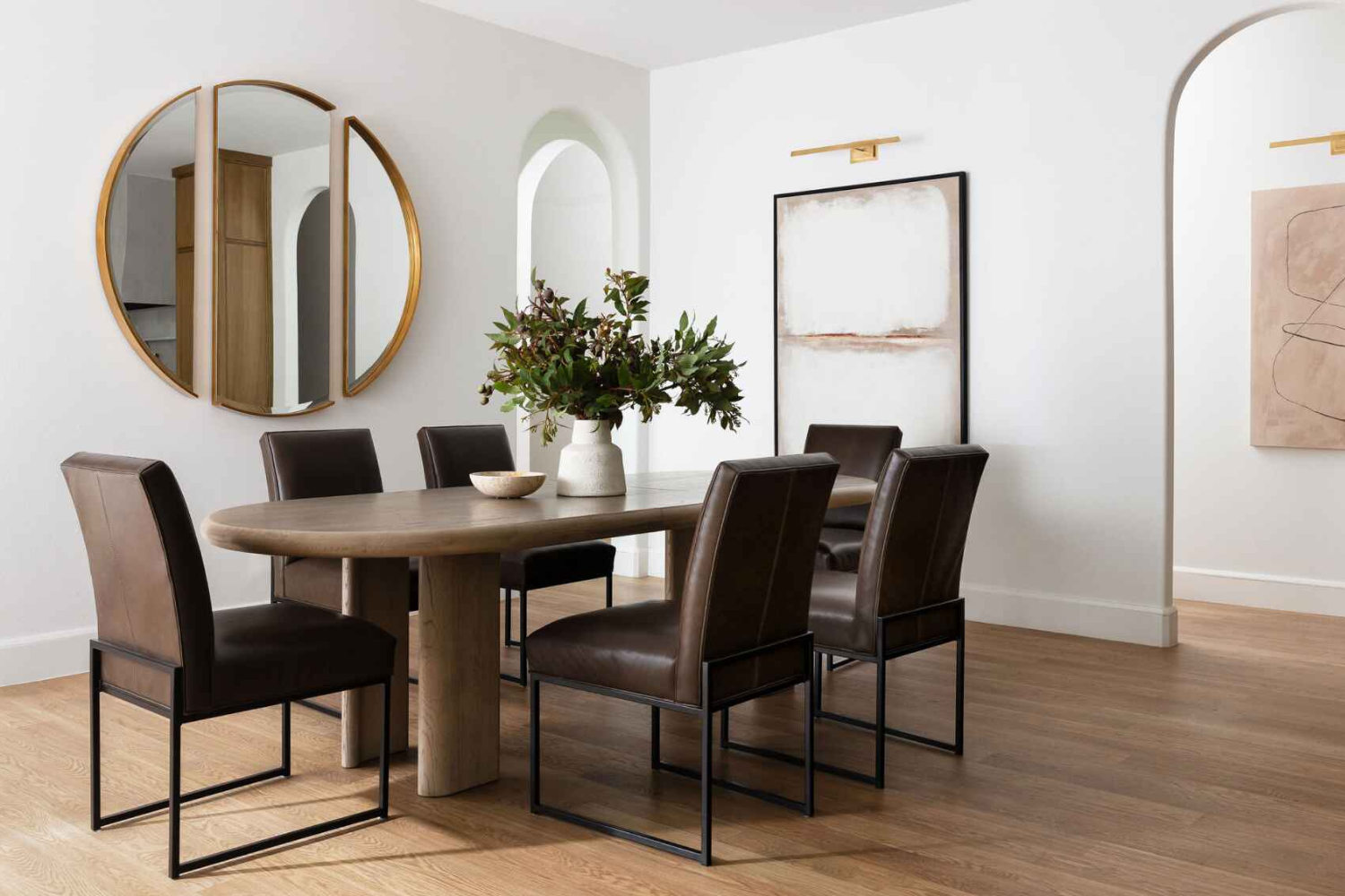 Transitional dining area with a sculptural wooden table, leather upholstered dining chairs, arched doorways, and minimalist abstract art.