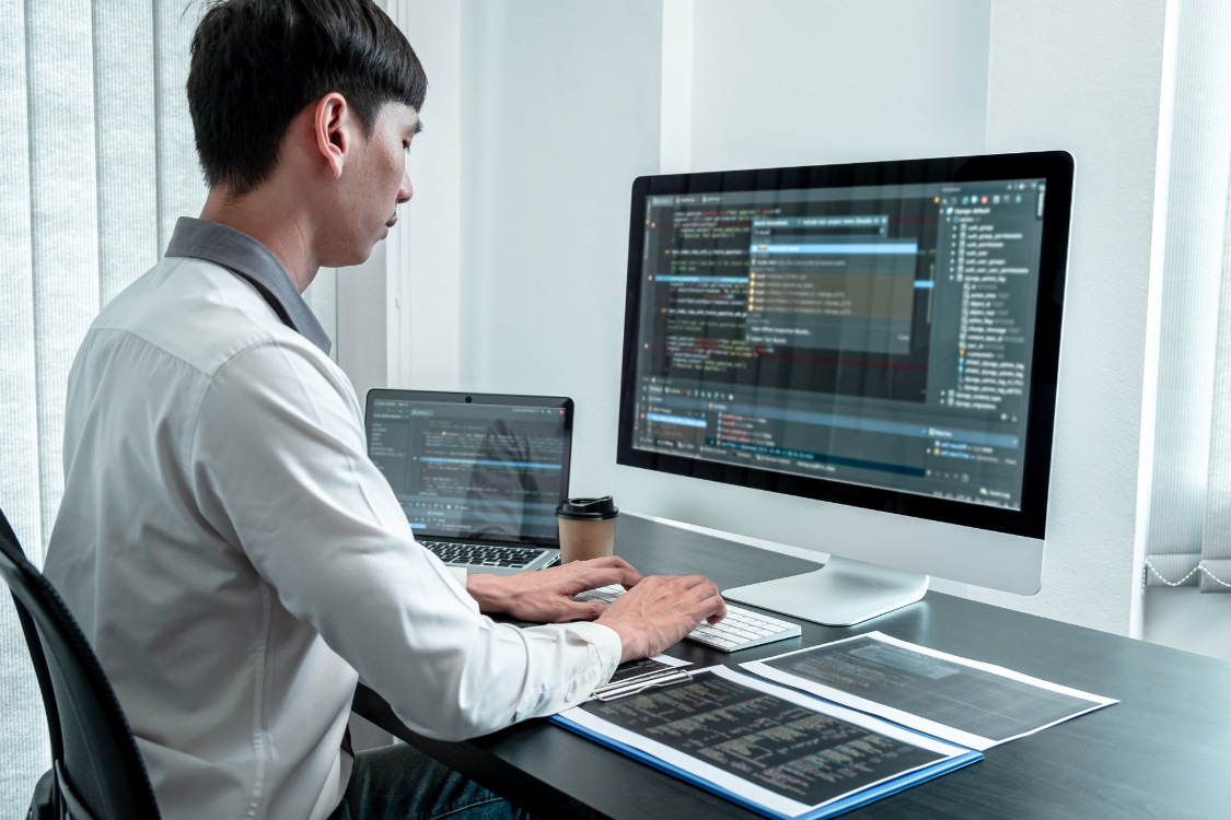 Filipino software developer working on code assessment with dual monitors and reference materials