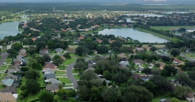 The image depicts a Mediterranean-style home exterior typical of the Orange Blossom Ranch development in Naples, FL, showcasing a spacious corner lot with lush landscaping and a tranquil residential community atmosphere. The home features an inviting entrance with French doors and a well-maintained yard, highlighting the luxurious dream house appeal of this gated community.