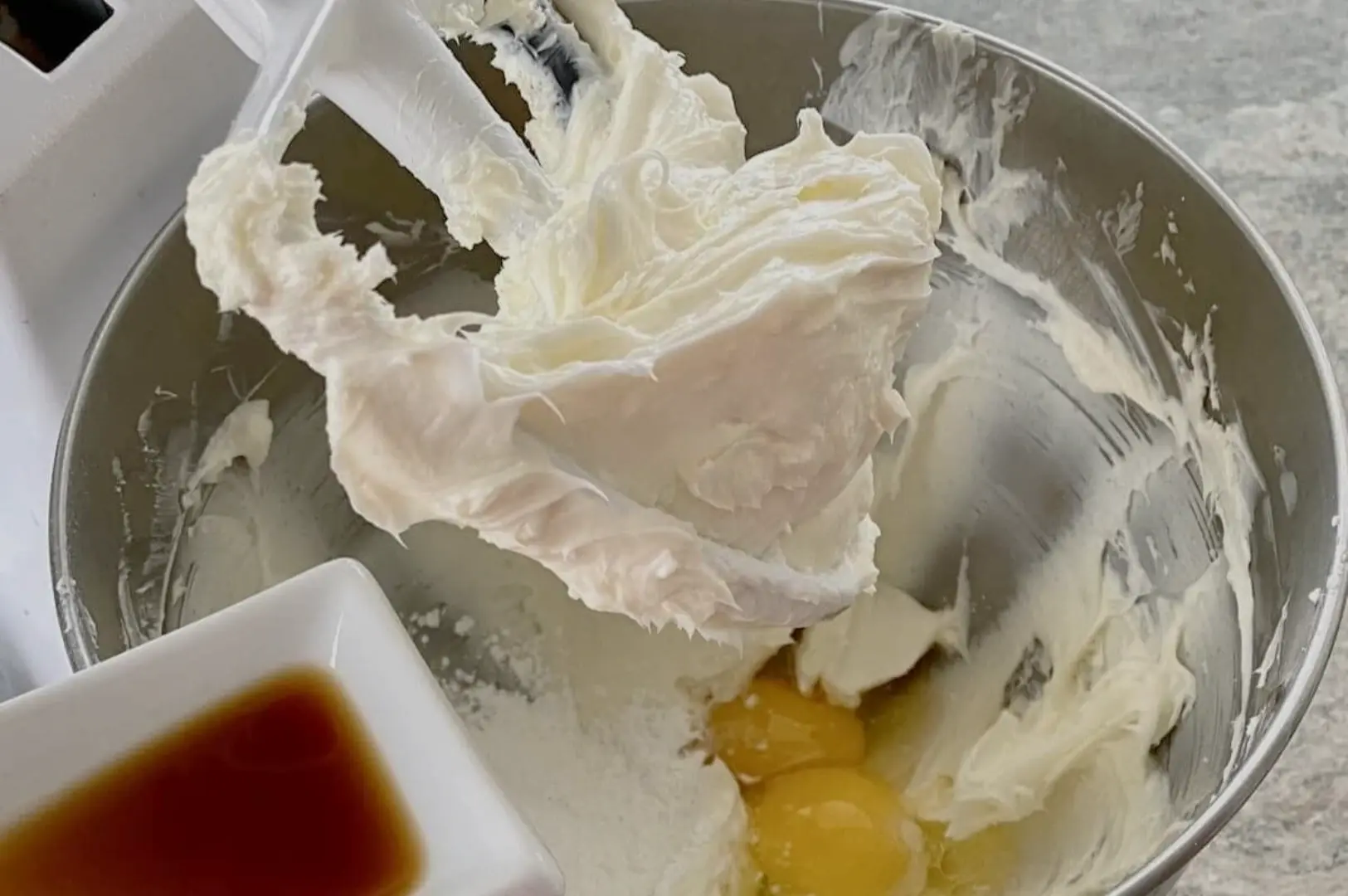 A mixer paddle lifts thick, fluffy white cream over a metal bowl with egg yolks and a small dish of brown vanilla nearby, ready for baking.