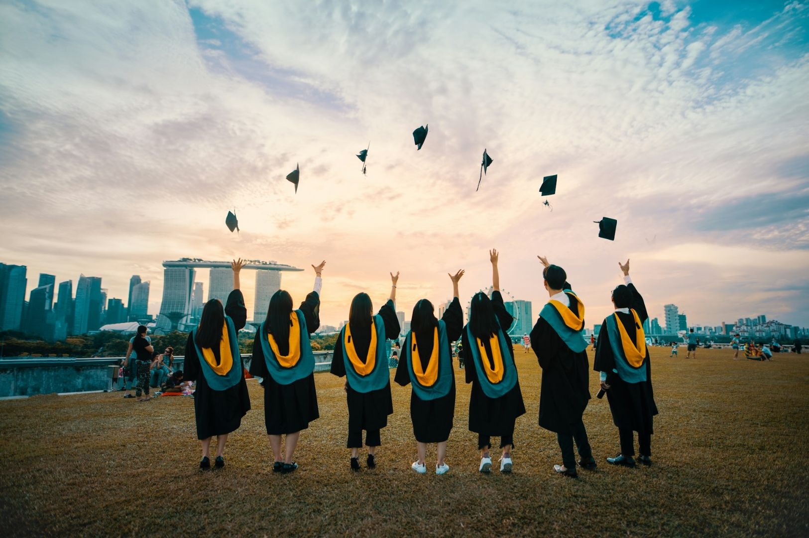Graduates in gowns and caps, viewed from behind, joyfully toss hats in the air. They stand on grass, with a city skyline and sunset in the background.