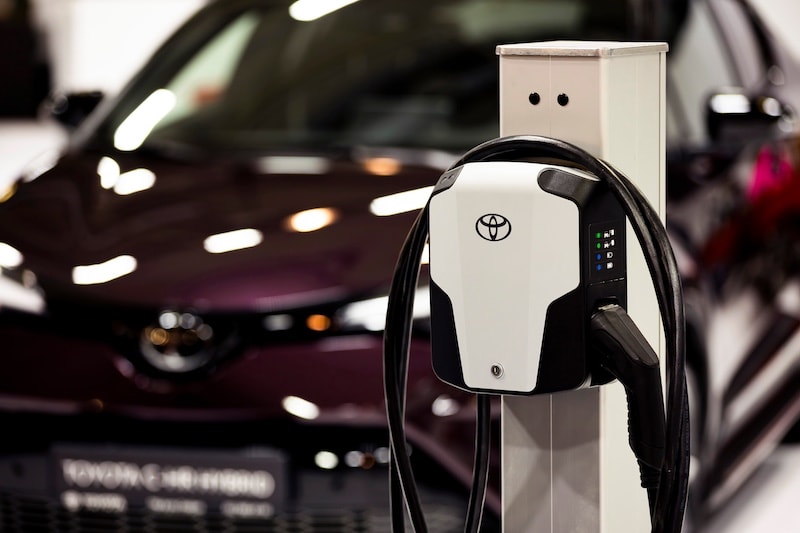 Close-up of a Toyota-branded electric vehicle charger with a hybrid car in the background, symbolizing the future of mobility at AutoFair Toyota of Tewksbury in Tewksbury, MA.