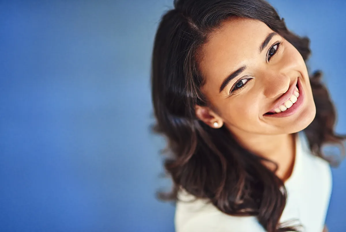 Portrait of a young woman with dark hair smiling against a blue background, showcasing her healthy white teeth.