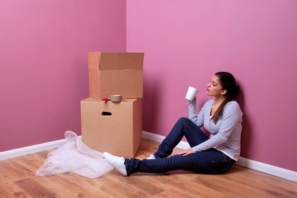 Girl sitting on the floor in a corner of the house, surrounded by boxes, holding a cup with a tired expression during a move.