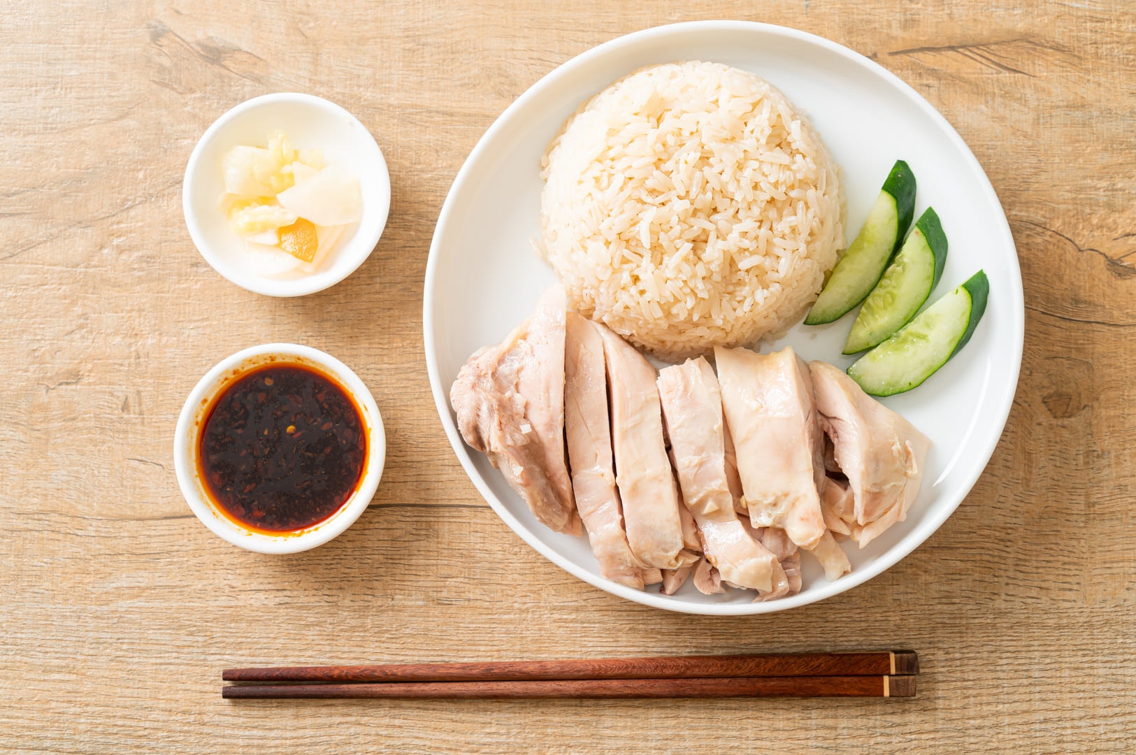 Hainanese chicken rice served on a plate with rice, tender chicken, cucumber slices, and chopsticks for eating.