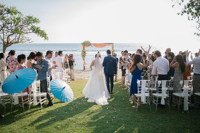 Bride walking accompanied to altar on beach with crowd watching on