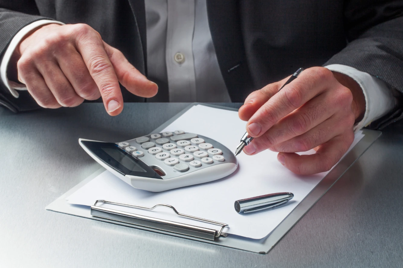 A person in a suit uses a pen and calculator on a clipboard.