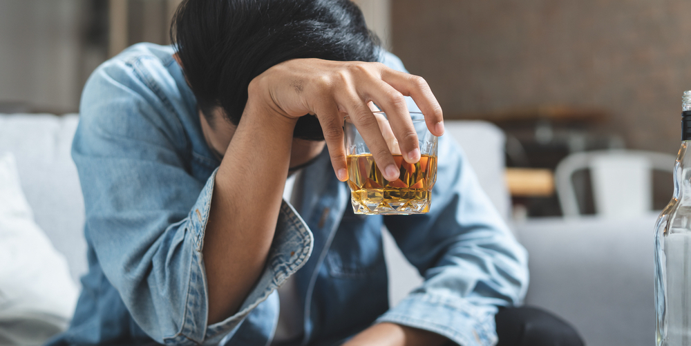 Man sits on the couch holding a half-full glass of whiskey, looking dejected and despairing