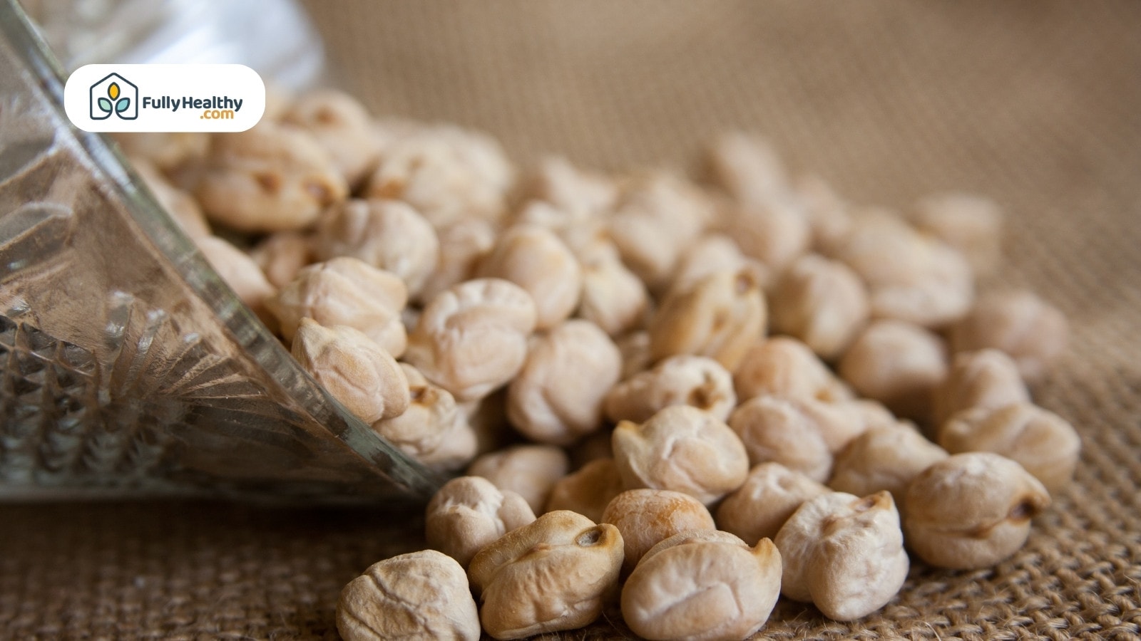 Raw chickpeas spilling from glass container onto burlap textured surface