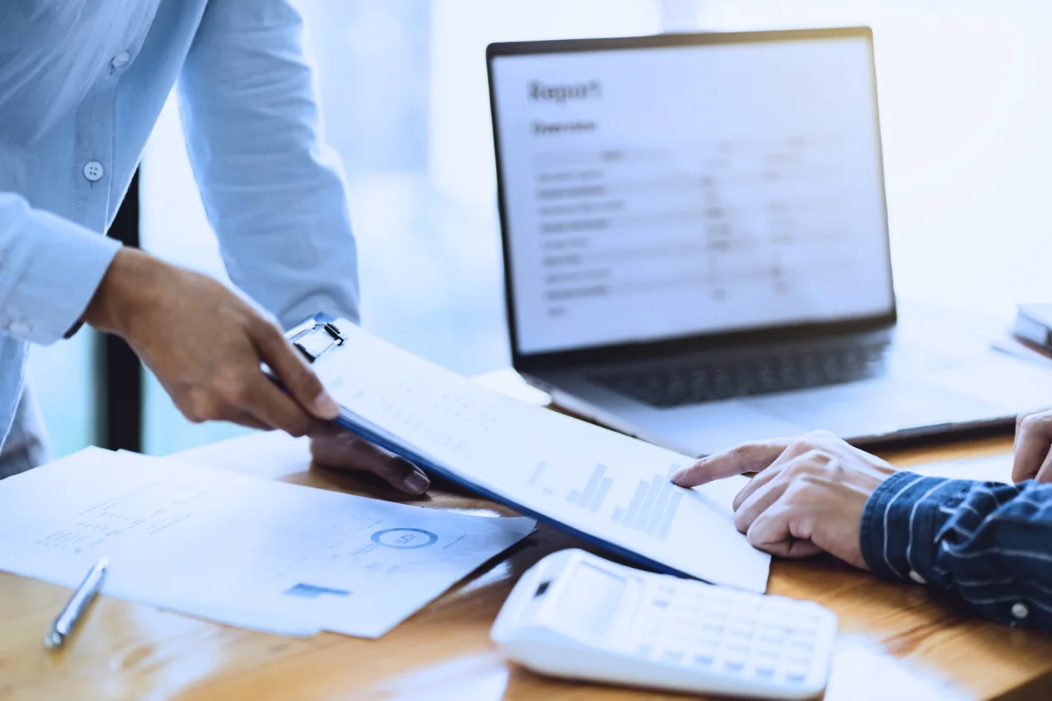 A person hands a document on a clipboard to another person who is pointing at it, with a laptop and calculator on the table. This reflects what is a valuation when selling a business.