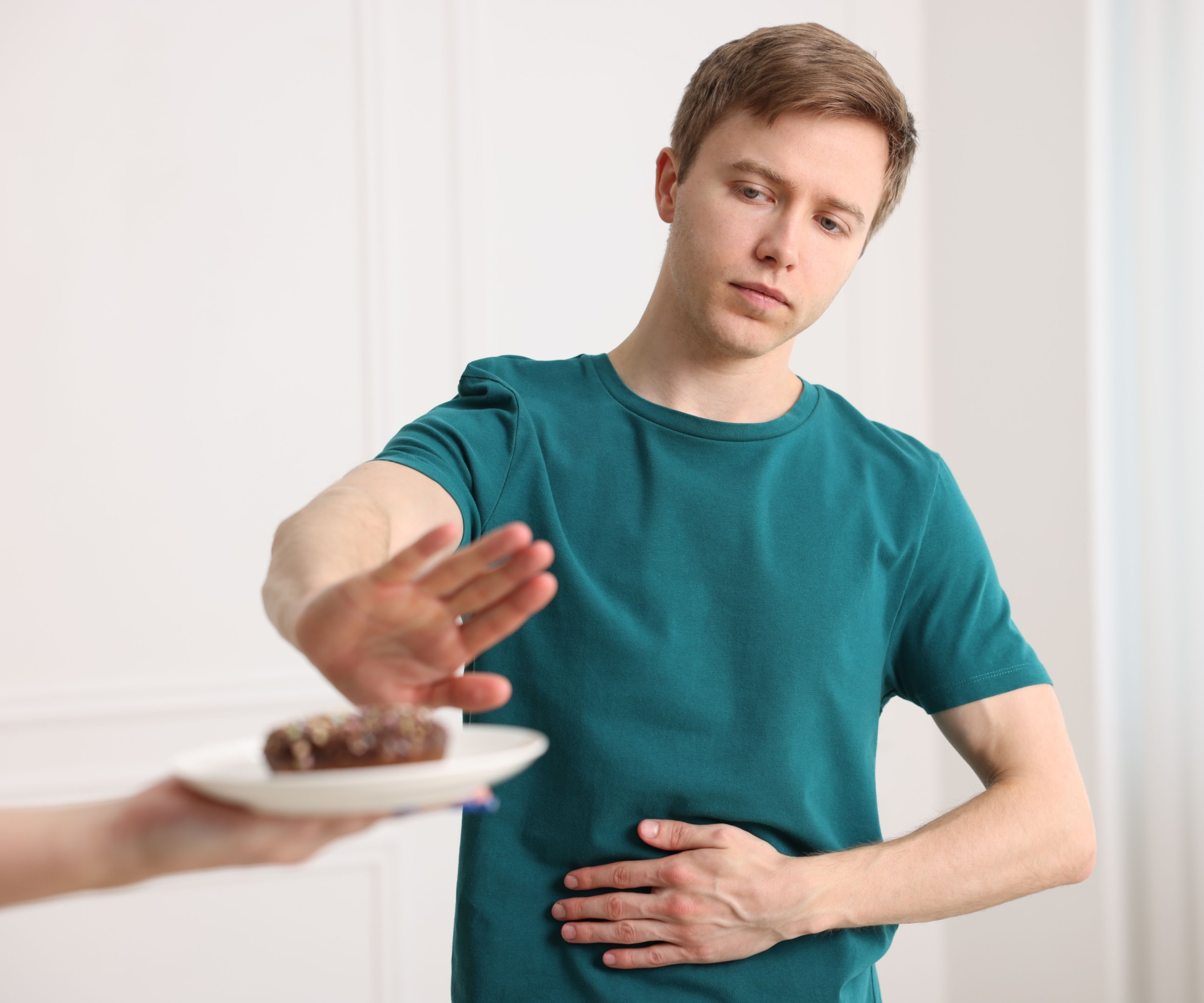 A man holding his stomach and gesturing a firm "stop" toward a plate of food, symbolizing the restrictive behaviors and physical discomfort often associated with eating disorders in men.