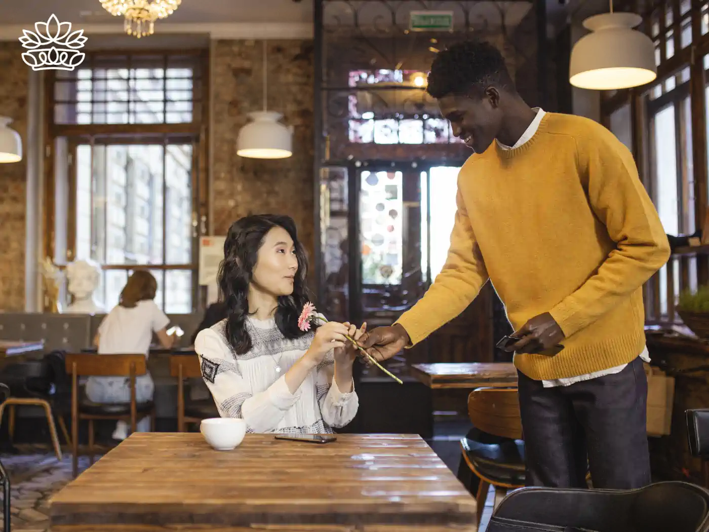 Young man in a yellow sweater presenting a delicate pink flower to a woman seated at a café table, capturing a moment of friendly exchange in a bustling coffee shop. Fabulous Flowers and Gifts - Thank You Flowers. Delivered with Heart