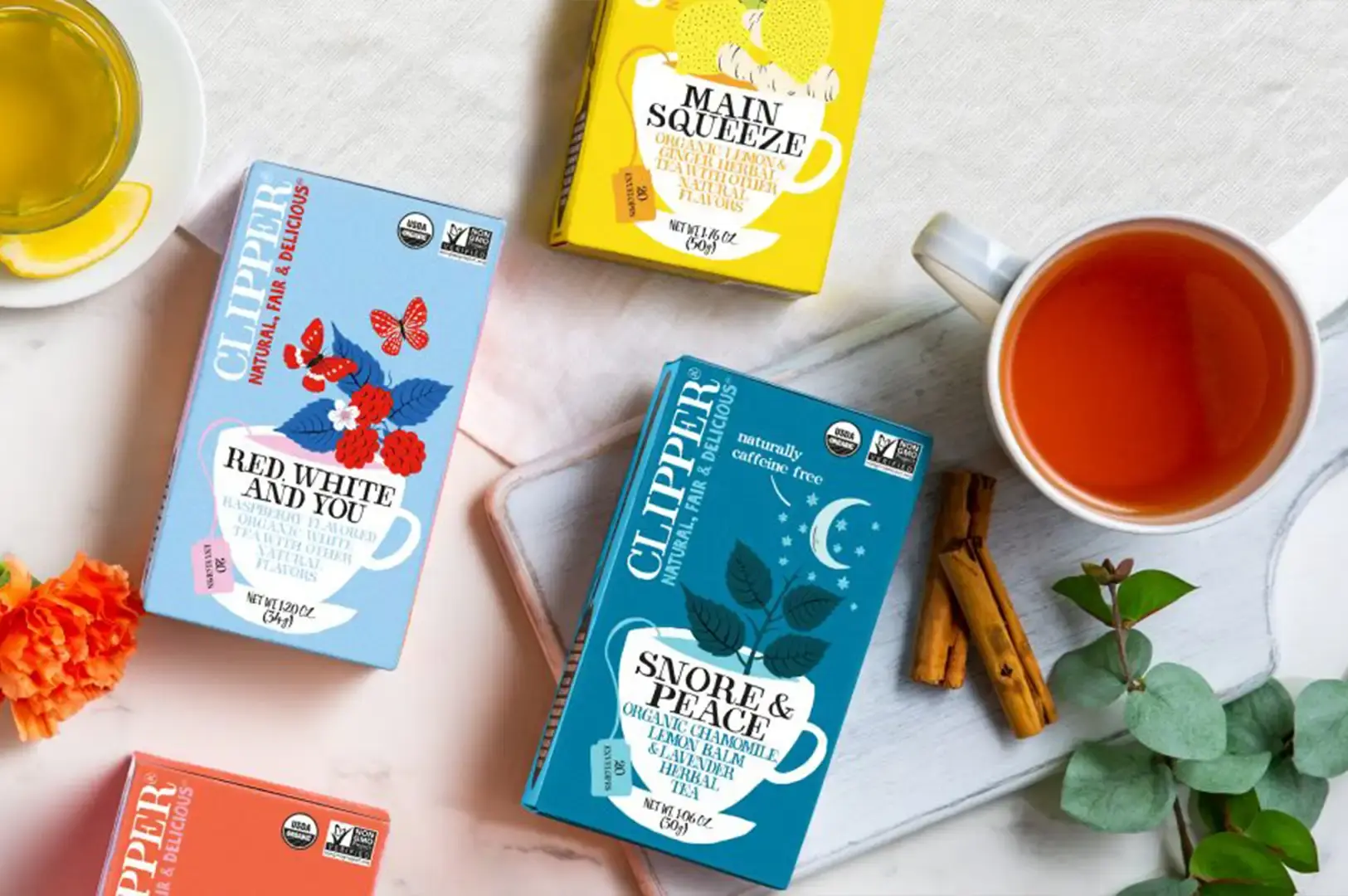 A top-down shot of various colorful Clipper tea boxes scattered across a light marble countertop alongside a glass mug of hot tea with a lemon slice. The boxes include "Red, White and You" white tea, "Main Squeeze" lemon and ginger tea, and "Snore & Peace" herbal tea, complemented by cinnamon sticks, fresh greenery, and a bright orange flower petal.