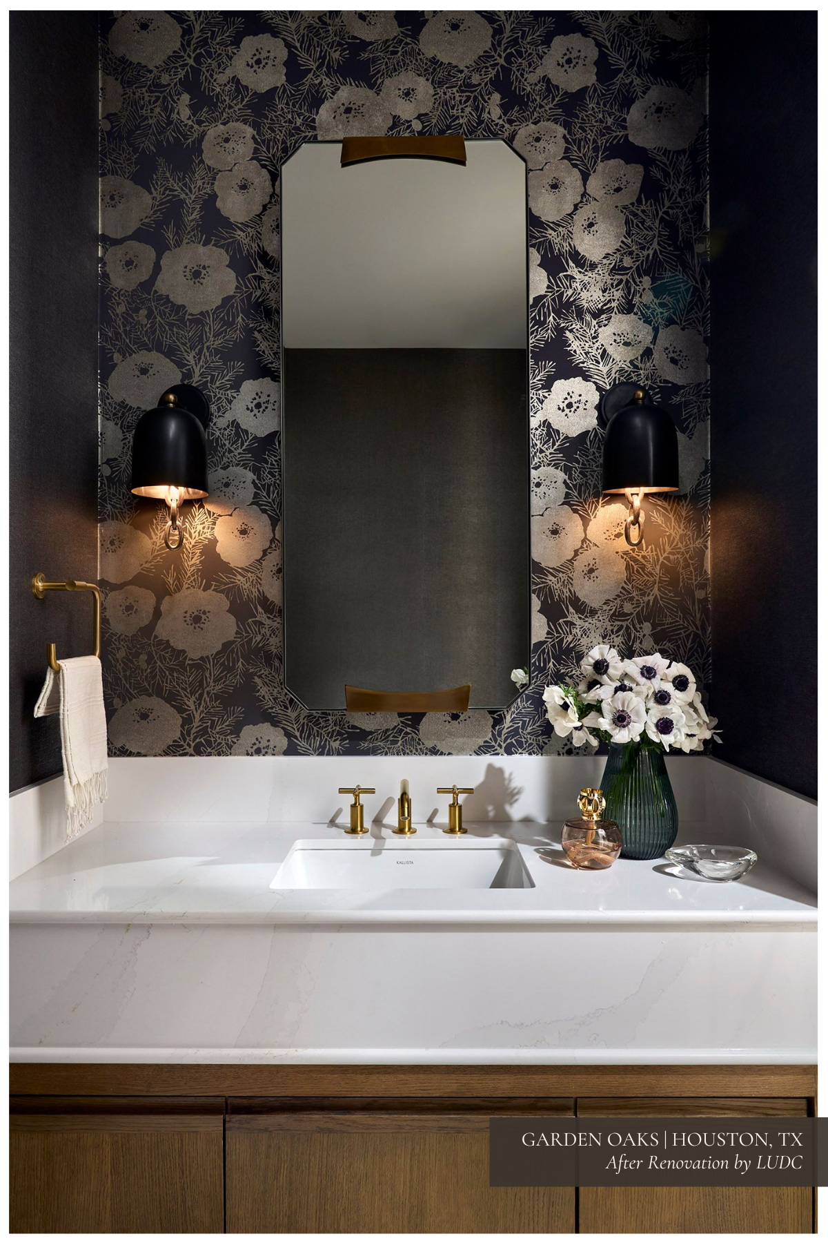 A luxurious powder room with a floral accent wallpaper by Romo, a gold-framed mirror, brass fixtures, and sconces by Arteriors, creating a sophisticated and dramatic ambiance.