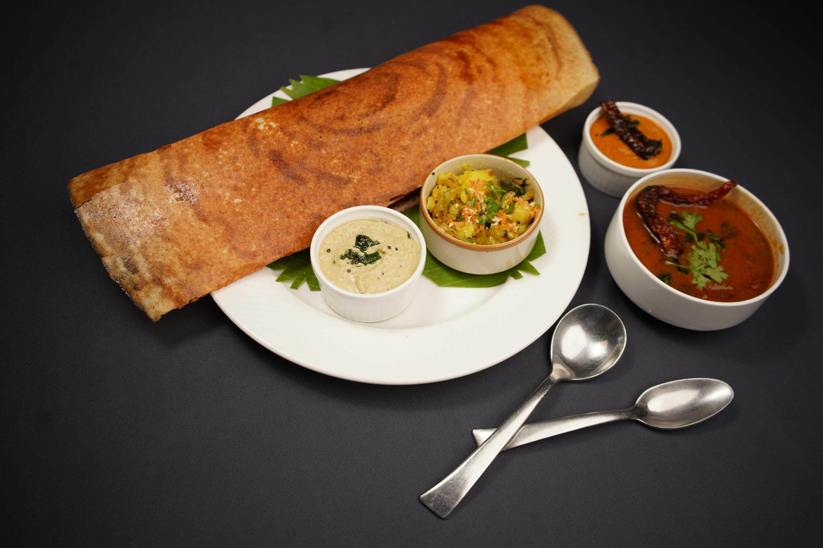 This image features a large, crispy golden-brown dosa rolled and served on a white plate with a green leaf garnish. Accompanying the dosa are four small bowls containing various chutneys, sambar, and a potato filling, along with two silver spoons set against a dark background.
