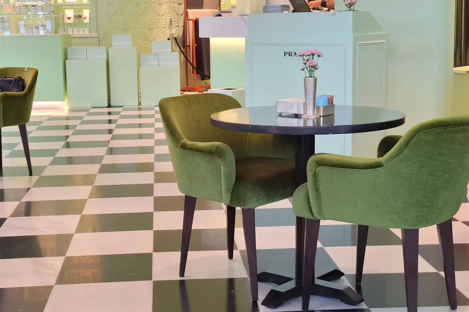 Dark green velvet armchairs around a small circular bistro table set on green and white checkered tiled flooring inside a Prada Cafe space.