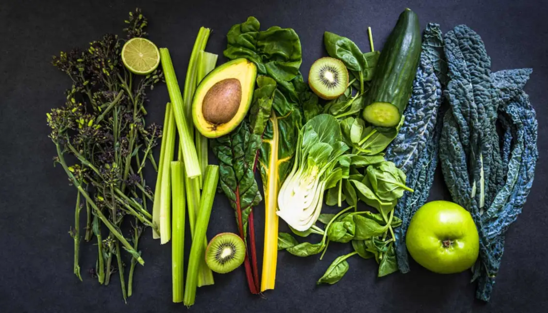 Grønne frukter og grønnsaker som avokado, kiwi, eple, selleri, spinat og grønnkål lagt pent utover en mørk bakgrunn."