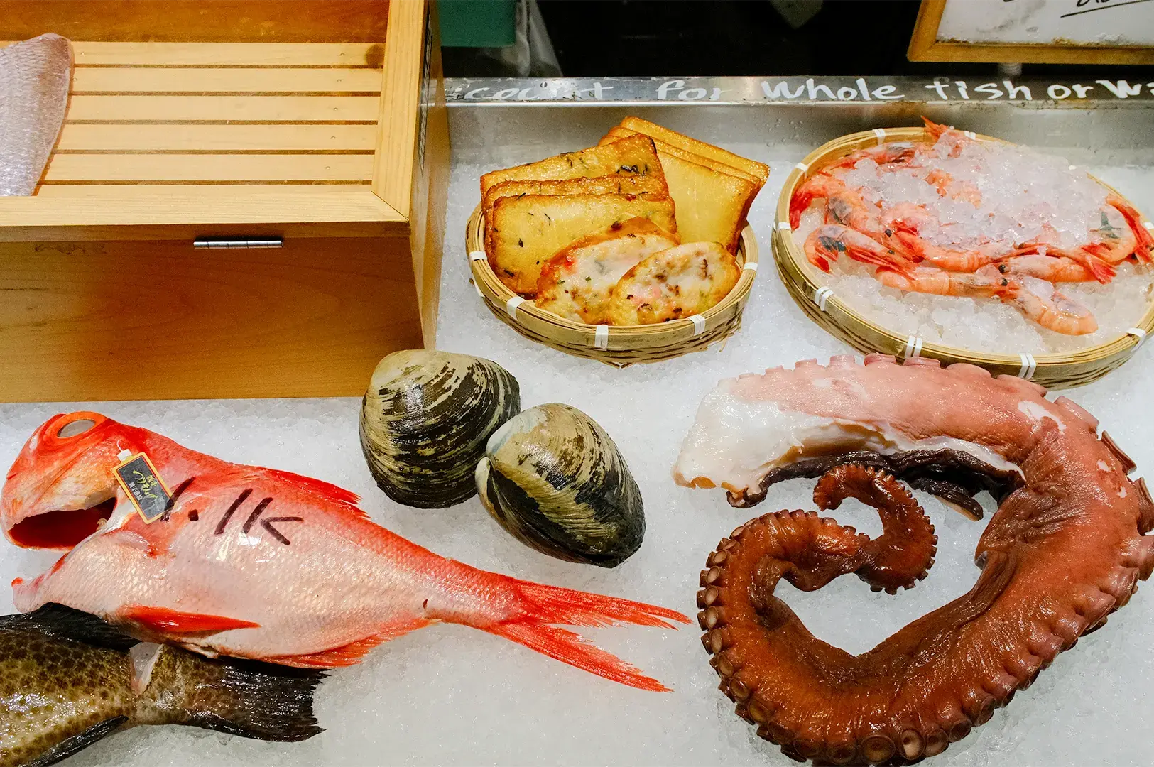 Assorted fresh seafood on ice featuring a red sea bream, giant octopus tentacle, large clams, sweet prawns, and fish cakes.