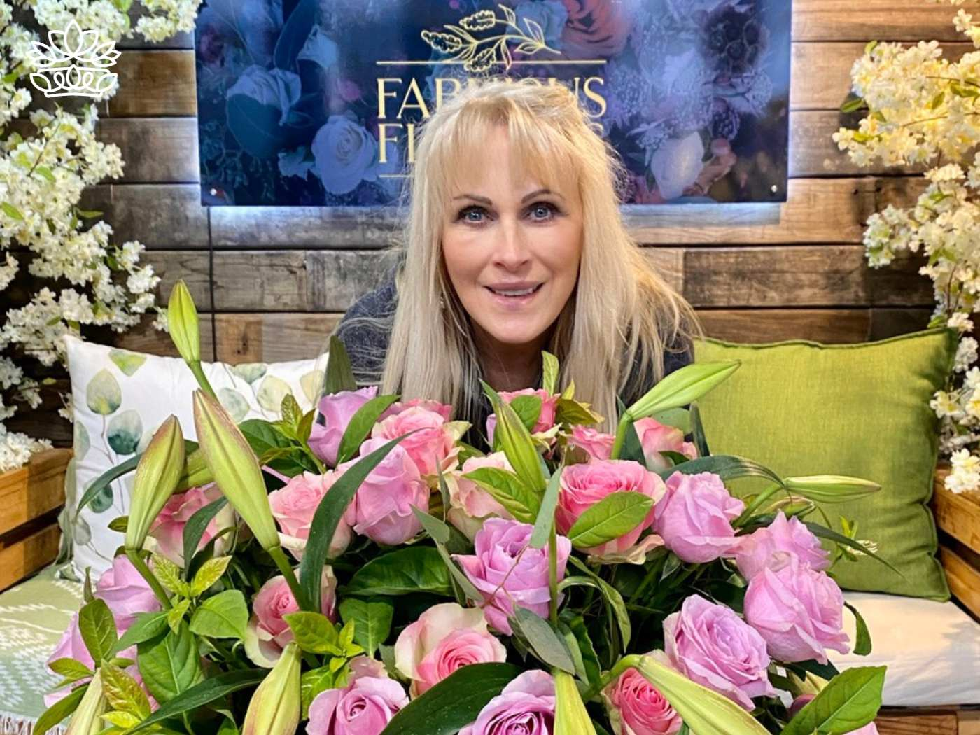 A woman with blonde hair smiles behind a vibrant display of pink roses and budding lilies, set in a cozy corner adorned with floral decorations at Fabulous Flowers and Gifts, part of the Flowers By Occasion Collection.