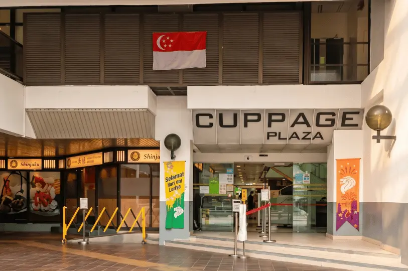 The exterior entrance of Cuppage Plaza in Singapore, featuring its name sign above glass doors, a hanging Singapore flag, and the storefront of Shinjuku Japanese restaurant to the left.